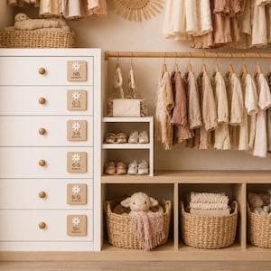 May include: A white six-drawer dresser with wooden knobs and square labels marked NB, 0-3, 3-6, 6-9, 9-12, and 12+ months. Beside it is a wooden closet organizer with hanging neutral-toned clothing, shelves for shoes, and three woven baskets containing plush toys.