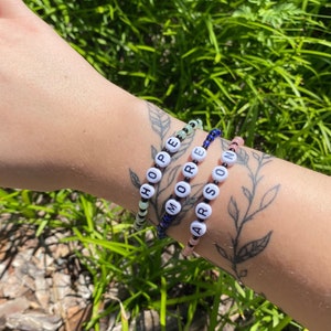 May include: Three beaded bracelets spelling out "HOPE", "MORE", and "ARSON" in white letter beads. The bracelets are strung on elastic and feature a variety of colored beads, including green, blue, and pink. The background is green grass.