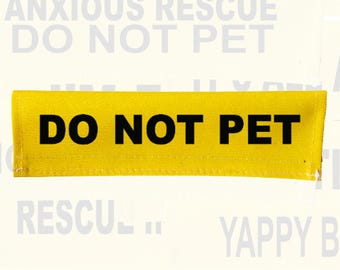 May include: A rectangular yellow fabric strap featuring the bold black text DO NOT PET centered on the front. The strap has visible stitching along the edges and is designed to be attached to a leash or harness for clear communication.