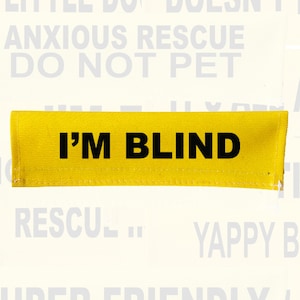 May include: A rectangular yellow fabric patch featuring the bold black text I'M BLIND centred on the front. The patch has stitched edges and is designed for high visibility, serving as a clear informational indicator for service animals.