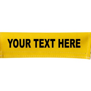 Op de afbeelding: Een gele stoffen band met zwarte stiksels en de tekst "YOUR TEXT HERE" in het zwart gedrukt.