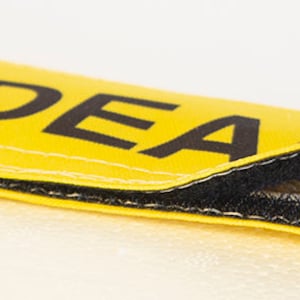 Op de afbeelding: Een gele stoffen band met zwarte stiksels en de tekst "I'M DEA" erop gedrukt. De band is gedeeltelijk opengezipt om een paars en gouden lint te laten zien.