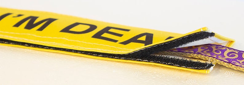Op de afbeelding: Een gele stoffen band met zwarte stiksels en de tekst "I'M DEA" erop gedrukt. De band is gedeeltelijk opengezipt om een paars en gouden lint te laten zien.