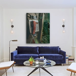 May include: A vintage, silver payphone with a red receiver hanging off the hook. The phone is mounted on a wall with green foliage behind it.