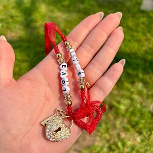 May include: A handmade charm with the word "LOVE" spelled out in white letter beads. The charm includes a gold heart-shaped lock with a key, a red heart, and gold and crystal beads, all attached to a red ribbon.