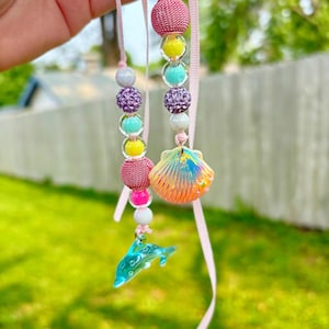 May include: A beaded necklace with a pink ribbon. The necklace features a variety of colorful beads, including pink, yellow, turquoise, and purple. A seashell pendant and a blue dolphin charm dangle from the necklace.