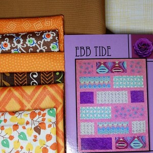 May include: A quilt pattern booklet with the title "Ebb Tide" on the cover. The cover is purple with a pink and white quilt pattern and a purple flower in the top right corner. The booklet is next to a stack of fabric scraps in orange, brown, and white.