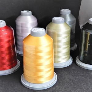 May include: Six spools of embroidery thread in various colours, including red, gold, beige, silver, and green. The spools are arranged in a semi-circle on a black surface.