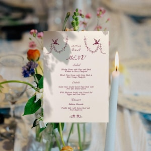 May include: A wedding menu card with a floral design and the text "MENU". The menu includes salad, entree, and dessert options. A lit white candle is in the background, with flowers in a vase.