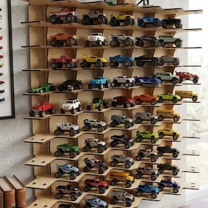 Puede incluir: Un estante de pared de madera con un diseño escalonado y dentado, que sostiene una gran colección de camiones monstruo y coches de juguete en miniatura. La estructura de madera contrachapada clara cuenta con múltiples niveles para organizar vehículos.