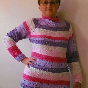May include: A striped sweater with a textured pattern in shades of pink, purple, and white. The sweater has a round neckline and long sleeves.