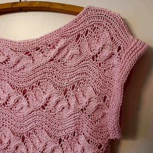 Puede incluir: Una blusa de crochet rosa con un delicado patrón de hojas. La blusa tiene un escote redondo y mangas cortas.