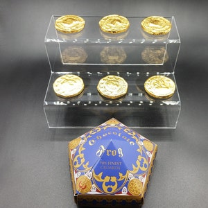 Może przedstawiać: Niebieskie i złote pudełko z napisem "Chocolate Frog" i "70% Finest Croakaoa". Pudełko ma kształt karty do gry i stoi na przezroczystym akrylowym stojaku z sześcioma złotymi czekoladowymi monetami.