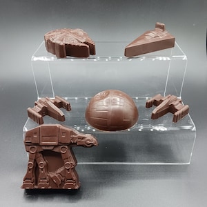 Puede incluir: Cinco figuritas de chocolate con temática de Star Wars en un expositor de acrílico transparente. Las figuritas son del Halcón Milenario, el caza TIE, la Estrella de la Muerte, el Ala-X y el AT-AT.