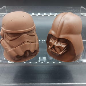 Puede incluir: Dos esculturas de chocolate de personajes de Star Wars, Darth Vader y un Stormtrooper, en un expositor transparente.
