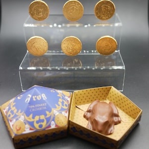 May include: Six gold chocolate coins on a clear stand. A chocolate frog sits in a star-patterned box, next to a closed box with the word "Frog" on it. The box is blue and gold.