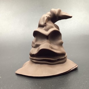 Peut inclure: Un chapeau parlant sculpté en brun avec un visage ridé et froncé. Le chapeau est un personnage populaire de la série Harry Potter.