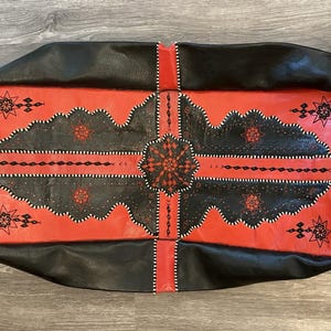 May include: A rectangular black and red leather bag with intricate geometric patterns. The bag features a central red panel with black decorative elements and star-shaped accents. The edges are black, and the design is stitched with white thread.