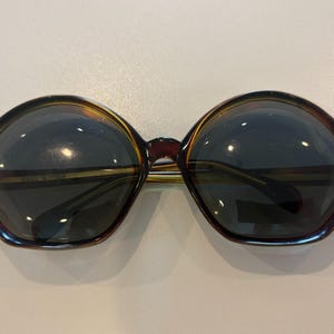 May include: Sunglasses with a dark brown tortoiseshell frame and large, rounded hexagonal lenses. The lenses are dark gray and provide full coverage. The frame features a smooth, polished finish with a bridge connecting the two lens sections.