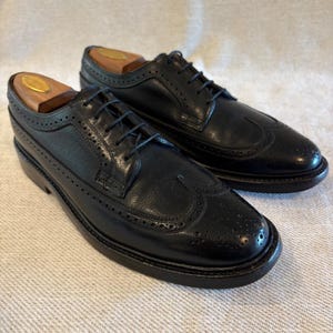 May include: A pair of black leather brogue shoes with wooden shoe trees. The shoes have a classic wingtip design with decorative perforations and a lace-up closure. The soles are dark brown.