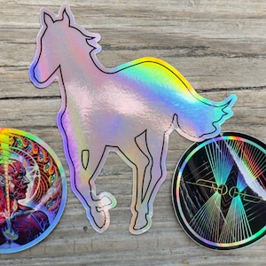 May include: Three holographic stickers on a wooden surface. The stickers include a horse silhouette, a circular design with a human figure, and a circular design with geometric lines. The stickers have a rainbow sheen.