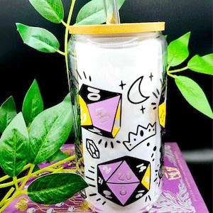 Puede incluir: Una lata reutilizable de vidrio con tapa de bambú y una pajita de vidrio. La lata tiene un fondo blanco con una ilustración en blanco y negro de dados, una corona, una luna creciente y cristales. Los dados son morados, amarillos y negros con números blancos.