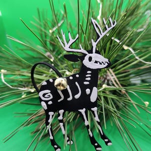 以下が含まれることがあります： 金色のベルが付いた黒と白の鹿の骨格のオーナメント。オーナメントは緑色の松の枝にぶら下がっています。