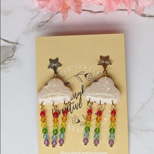 May include: Cloud-shaped earrings with rainbow bead accents. The earrings feature a white cloud design with gold star accents at the top. The rainbow beads cascade downwards, creating a colorful and whimsical look. The earrings are on a tan card.
