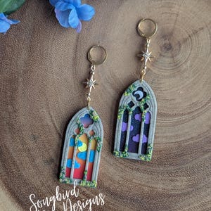May include: A pair of dangle earrings featuring a gothic window design. One earring depicts a sunset scene with a moon and clouds, while the other shows a night sky with purple clouds. The earrings are adorned with gold-colored hoops and star accents. The text "Songbird Creative Designs" is visible.