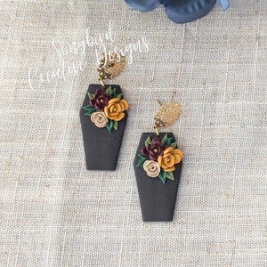 May include: A pair of black coffin-shaped earrings with gold accents and floral arrangements. The earrings feature a cluster of burgundy, mustard yellow, and cream-colored flowers. The text "Songbird Creative Designs" is visible.