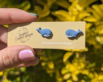 May include: A pair of blue whale stud earrings on a tan card. The earrings are small, with a light blue body and a white belly. The card has the text "Songbird Creative Designs" and the Instagram handle @songbirdcreativedesigns.