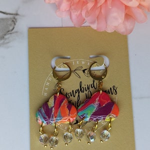 May include: Colorful cloud-shaped earrings with gold-colored hoops and clear crystal bead dangles. The earrings feature a marbled design in shades of red, orange, purple, and green. The earrings are displayed on a tan card with text.