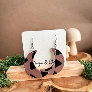 May include: A pair of brown and black crescent moon shaped earrings with a black and white geometric pattern. The earrings are on a white card with the text "Onyx & Clay".