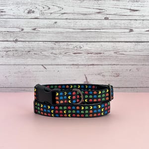 May include: Black dog collar featuring a colorful pattern of video game characters. The collar has a black plastic buckle and a metal D-ring for leash attachment. The background is a light pink surface with a white wooden backdrop.