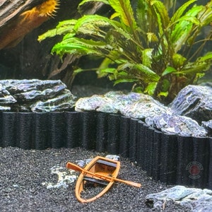 May include: A small wooden boat with oars rests on a bed of black gravel in a fish tank. The tank is decorated with rocks and a green plant.