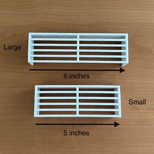 K&ouml;nnte beinhalten: Zwei wei&szlig;e, rechteckige L&uuml;ftungsgitter, eines mit der Aufschrift "Large" und einer Gr&ouml;&szlig;e von 15 cm, das andere mit der Aufschrift "Small" und einer Gr&ouml;&szlig;e von 13 cm. Beide haben horizontale Lamellen und sind auf einer braunen Oberfl&auml;che dargestellt.