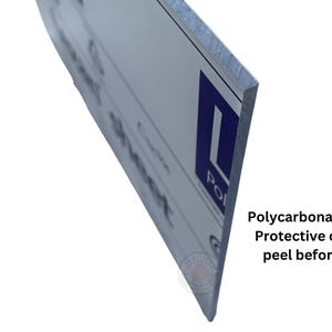 May include: Close-up of the edge of a white sign with blue text. The sign is made of 3/16" polycarbonate with a protective coating that needs to be peeled off before use.