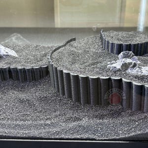 May include: Three black plastic platforms with a textured, corrugated edge, arranged in a tiered formation. The platforms are filled with black sand and feature small, gray rocks.