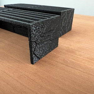 K&ouml;nnte beinhalten: Schwarze, 3D-gedruckte Erh&ouml;hungen mit einem strukturierten, geometrischen Design. Die Erh&ouml;hungen haben eine Lamellenoberseite und sind so konzipiert, dass sie Objekte anheben. Das Design weist eine Reihe horizontaler Linien auf der Oberseite und ein strukturiertes, eckiges Muster an den Seiten auf.