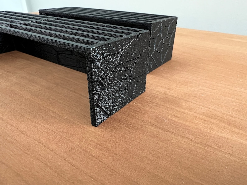 K&ouml;nnte beinhalten: Schwarze, 3D-gedruckte Erh&ouml;hungen mit einem strukturierten, geometrischen Design. Die Erh&ouml;hungen haben eine Lamellenoberseite und sind so konzipiert, dass sie Objekte anheben. Das Design weist eine Reihe horizontaler Linien auf der Oberseite und ein strukturiertes, eckiges Muster an den Seiten auf.