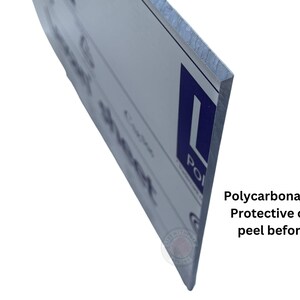 May include: Close-up of the edge of a white sign with blue text. The sign is made of 3/16" polycarbonate with a protective coating that needs to be peeled off before use.