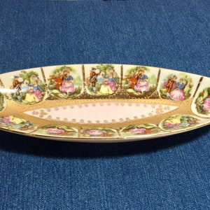 May include: An oval-shaped serving dish with a pink and gold design. The dish is decorated with a repeating pattern of couples in a garden setting.