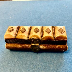 May include: A rectangular wooden box with a hinged lid. The box is primarily brown with cream-colored accents and decorative diamond-shaped embellishments. The box has a brass clasp and hinges. The box is on a blue surface.