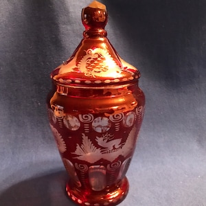 May include: A red glass jar with a lid, decorated with white etched designs of birds and swirls. The jar has a rounded body and a wide mouth.