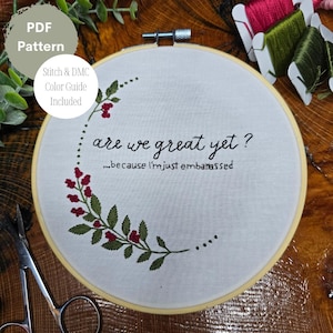 May include: Embroidered hoop art with a white fabric background and a green leafy branch with red berries. The text reads "are we great yet? ...because I'm just embarrassed".