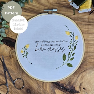 May include: A white embroidery hoop with a quote that reads "Some of those that hold office are the same that burn crosses." The quote is embroidered in black thread. The hoop is decorated with yellow flowers and green leaves embroidered in thread.