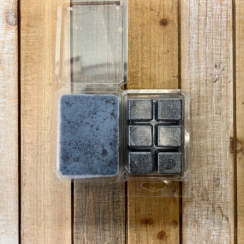 May include: Two clear plastic containers filled with black wax melts. One container holds a single large melt, while the other holds six smaller square melts.