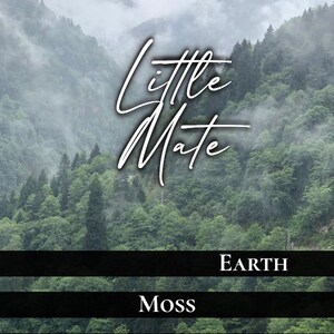 May include: A candle label with the text "Little Mate" in a cursive font, set against a backdrop of a misty mountain forest. The label includes the words "Earth," "Moss," and "Orc Dick Magic."