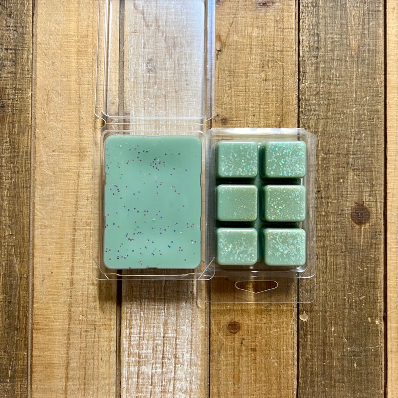 Puede incluir: Dos recipientes de pl&aacute;stico transparente con ceras arom&aacute;ticas de color verde salvia. Un recipiente est&aacute; abierto, revelando un bloque rectangular s&oacute;lido, mientras que el otro est&aacute; cerrado, mostrando seis cubos de cera cuadrados. Ambos tienen purpurina iridiscente.