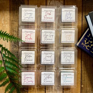 May include: Twelve small square containers of wax melts, each with a different scent and name. The names are all romance-themed, such as "Grumpy Sunshine", "Dark Romance", "Why Choose", "Smut Reader", "Age Gap Romance", "Single Dad", "Mafia Romance", "Forced Proximity", "Morally Grey", "Enemies to Lovers", "Stalker Romance", and "Billionaire Romance".